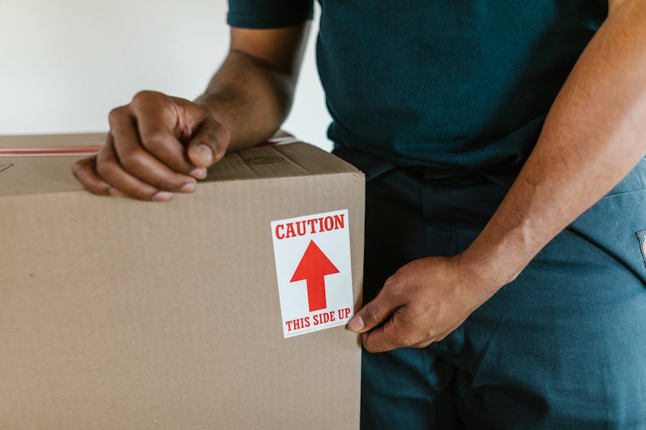 Close-up of a person wearing a dark green shirt and blue pants, holding a cardboard box with both hands. The box has a white and red caution label that reads 'CAUTION' with an upward arrow and 'THIS S
