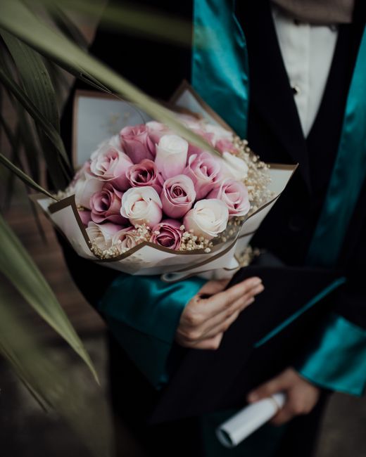 A person dressed in a teal satin blazer and white shirt is holding a bouquet of fresh pink and cream roses, arranged in a cluster with small white filler flowers, wrapped in white and beige paper. The