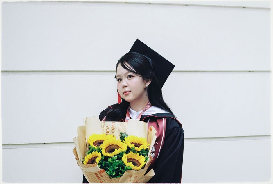 A young woman dressed in a black graduation gown with a red and white hood, wearing a traditional mortarboard cap, holds a bouquet of bright yellow sunflowers wrapped in brown paper with red accents, 