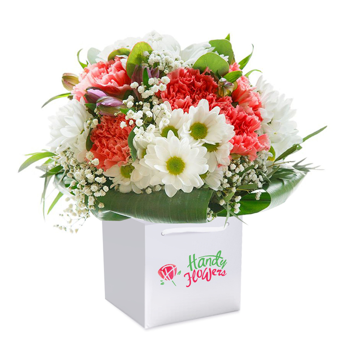 Displayed tall and proud within a sleek, square white gift bag, this Roehampton bouquet is a celebration of harmonious hues and refined local floristry. Coral-orange, peach, and deep red carnations-in all their frilly splendor-form the central cluster, their softness balanced by the crisp edges and radiant centers of white daisies reminiscent of blooms in Richmond Park's wildflower walks. Baby's breath, with each tiny white blossom, adds a delicate, airy texture, softening the bolder florals and lending the arrangement a dreamy touch. Here and there, purple-tinged buds still tightly closed introduce a note of mystery and depth. Anchoring the composition is a generous sweep of glossy, deep green foliage, the broad leaves enfolding the bouquet in a naturally elegant collar. The result is a sensuous, rounded display exuding fullness and vitality. The white gift bag, fitted with robust ribbon handles, offers practicality for delivery from the very heart of Roehampton to the doorstep of a loved one, a local business, or a friend near the University campus. Against a seamless white background, every flower stands out in clear relief, emphasizing freshness and expert arrangement. Perfect for celebrations, heartfelt thanks, or simply to bring a splash of Roehampton's cheer into any space.