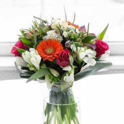 Combining timeless style with Roehampton's leafy heritage, this splendid bouquet captures the essence of fresh beginnings and heartfelt celebration. A quartet of gerbera daisies-two vibrant orange, two in creamy-peach-stand out at the bouquet's centre, their velvety petals adding a lively surge of colour. Interwoven among them are rich magenta roses, whose deep, tightly curled buds lend a sumptuous, romantic note. Abundant sprays of white alstroemeria, alongside petite filler blooms in soft pink and snowy white, infuse a lightness that conjures images of early summer along Roehampton's peaceful avenues. An impressive base of glossy, dark green leaves, shaped into broad, elegant curves, grounds the design-mirroring the structured yet natural beauty found in the neighbourhood's gardens and open spaces. Slender green sprigs punctuate the arrangement, filling every gap and lending a layered, finished look. The clear glass vase accentuates the stems' vibrant green, reinforcing the bouquet's sense of freshness and local pride. Illuminated by gentle daylight, the composition feels both luxurious and approachable, perfect for any special occasion or to bring a touch of the outdoors into a Roehampton home. This expertly arranged bouquet, available for local delivery, carries with it the scents and colours of the season, making every recipient in SW15 feel remembered and cherished.